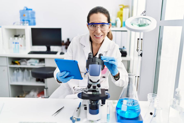 Jonge Spaanse vrouw draagt wetenschapper uniform met touchpad en microscoop in het laboratorium - Foto, afbeelding