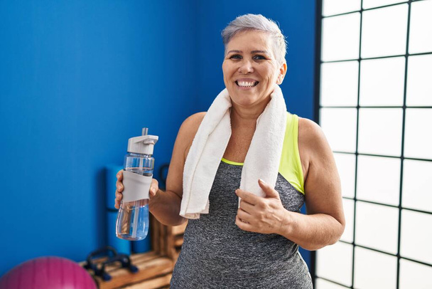 Middle age woman wearing sportswear and towel holding water looking positive and happy standing and smiling with a confident smile showing teeth  - Фото, зображення