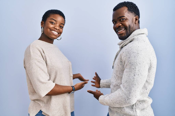Молодая африканская американская пара, стоящая на синем фоне вместе, приглашает к естественной улыбке с открытой рукой  - Фото, изображение