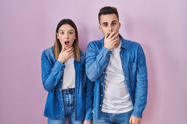 Young hispanic couple standing over pink background looking fascinated with disbelief, surprise and amazed expression with hands on chin  - Zdjęcie, obraz