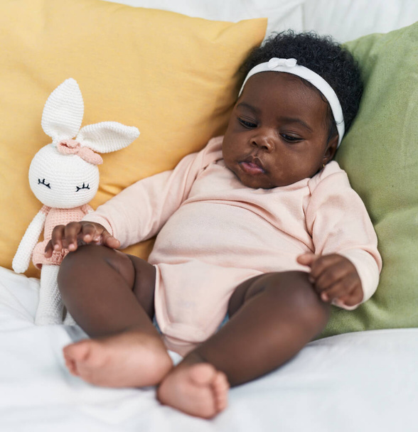 Bebé afroamericano sentado en la cama con expresión relajada en el dormitorio - Foto, imagen