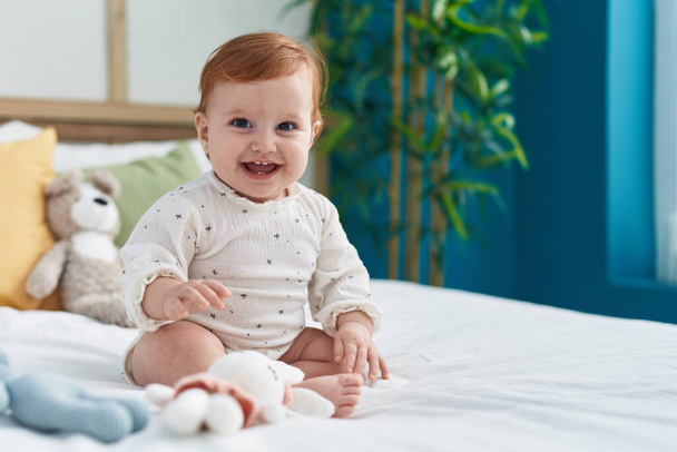 Imádnivaló vörös hajú kisbaba mosolyogva magabiztos ül az ágyon a hálószobában - Fotó, kép