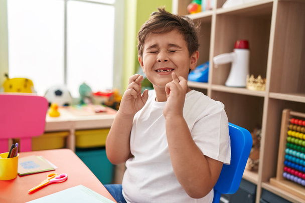 Imádnivaló spanyol óvodás fiú, aki az asztalnál ül és keresztbe teszi az ujjait az óvodában. - Fotó, kép