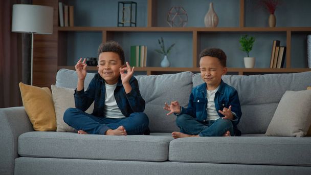 Two funny little African American ethnic children boys brothers siblings kids meditating together sitting on couch in lotus position. Small schoolboys meditate. Spiritual development yoga meditation - Foto, Imagem