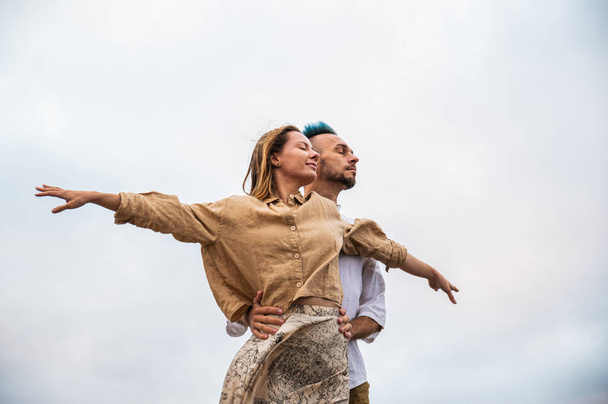 A young couple stands with their arms open and eyes gazing into the distance on a rocky cliff by the ocean. The man has his hands on the womans waist, and she has her arms outstretched. The couple - Foto, afbeelding