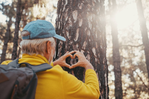 Head shot portrait close up of one old cute man taking care and protecting big tree in the forest of mountain in the nature. One mature person making a heart shape with hands loving nature concept. - Foto, imagen