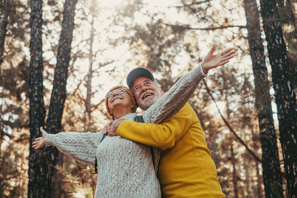 Head shot portrait close up of middle age cheerful people smiling and looking at the the trees of the forest around them. Active old woman opening arms with husband hugging her from back - Fotografie, Obrázek