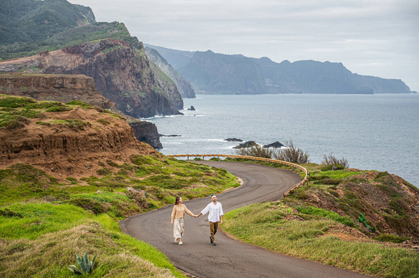 A distance shot of a young couple walking on a winding road, holding hands. The road is surrounded by lush green grass, cliffs and ocean. The couple appears to be taking in the natural beauty of the - Valokuva, kuva