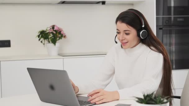 Young woman in headset conducts online meeting with team via Notebook. Manager discusses details of new project sitting at table slow motion - Imágenes, Vídeo