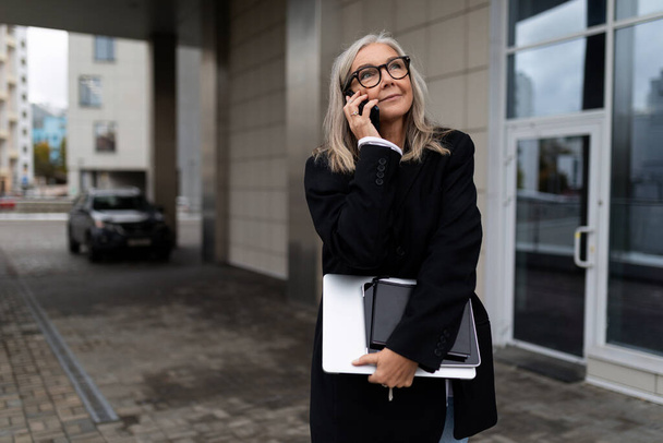 femme mûre avec un ordinateur portable dans ses mains parlant sur un téléphone portable à côté de l'entrée de l'immeuble de bureaux. - Photo, image