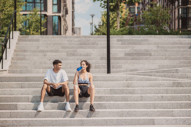 Молодая женщина сидит на бетонных ступеньках и пьет воду в присутствии своего веселого партнера по фитнесу - Фото, изображение