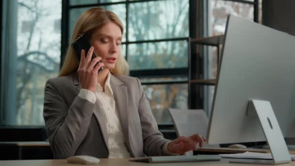 Caucasian middle-aged adult woman with computer talk mobile phone discuss deal business consultation. Female 30s businesswoman worker employer in corporate office talking with smartphone multitasking - Filmmaterial, Video