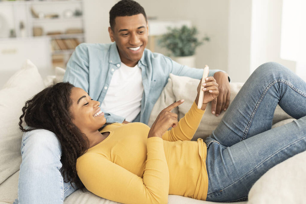 笑顔の若い黒のカップルは、スマートフォンでビデオを見て、リビングルームのインテリアでソファの上で自由な時間に一緒に休息とリラックスをお楽しみください。新しいアプリ,ソーシャルネットワークでチャット,ライフスタイル,広告や自宅で提供 - 写真・画像