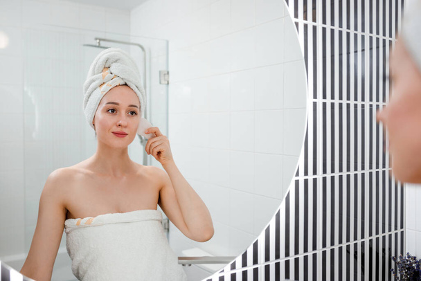 Mladá krásná žena v bílém ručníku dělá masáž obličeje s růžovým křemenem scraper během krásy ranní rutiny v koupelně. Anti aging léčby a péče o pleť koncept - Fotografie, Obrázek