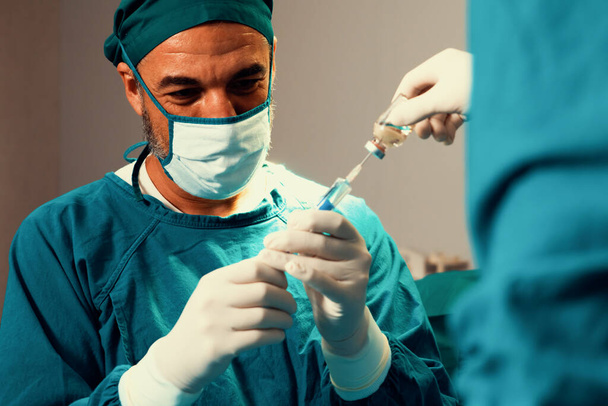 Kirurgi täyttää ruiskun lääketieteellisestä injektiopullosta kirurgiseen toimenpiteeseen steriilissä leikkaussalissa avustavan sairaanhoitajan kanssa. Lääkäri ja hoitohenkilökunta täysin suojaava kulumista leikkaukseen valmistella anestesia injektio - Valokuva, kuva