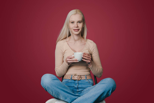 Beautiful positive woman sitting on chair holding cup of coffee and smiling at camera isolated over crimson background. Attractive beautiful girl. People - Foto, afbeelding