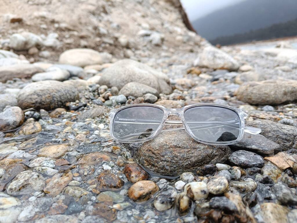 A pair of modern, stylish, and trendy sunglasses or shades or sunnies placed on the top of a rock in a stream of water with beautiful natural background. The sunglasses have grey tinted lenses - Foto, Imagem