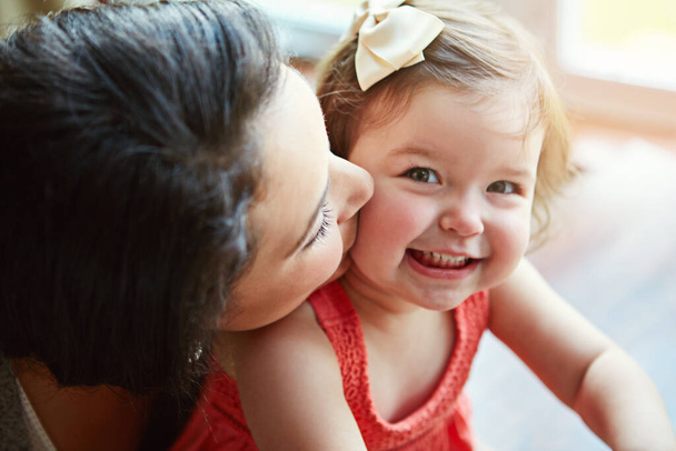 Portrait of a baby with her mother kissing her cheek while playing, bonding and spending time together. Happy, smile and girl infant child sitting with her mom in their modern family home with love - Foto, Imagen