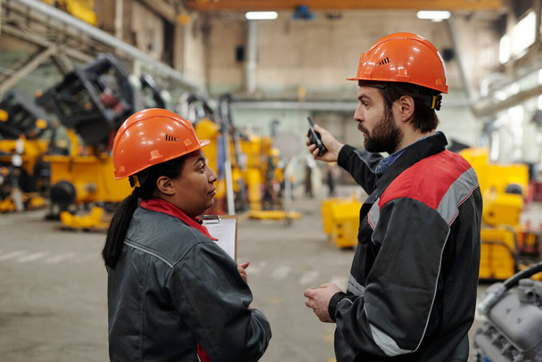 Two young confident interracial engineers in workwear having discussion of working points while standing in distribution warehouse - Fotografie, Obrázek