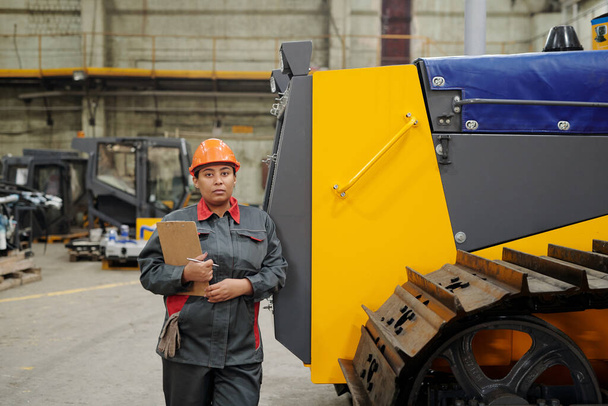 Young confident staff of large modern factory standing by huge industrial machine while working in warehouse - Foto, imagen