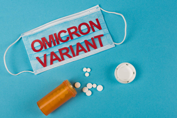 Top view of medical mask with omicron variant lettering near pills on blue background  - Foto, Imagen