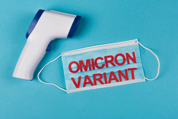 Top view of non-contact pyrometer near medical mask with omicron variant lettering on blue background  - Foto, Imagem