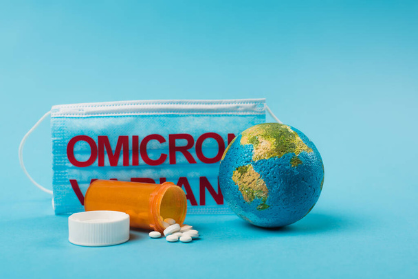 Глобус и таблетки рядом с медицинской маской с омикронным вариантом надписей на синем фоне  - Фото, изображение