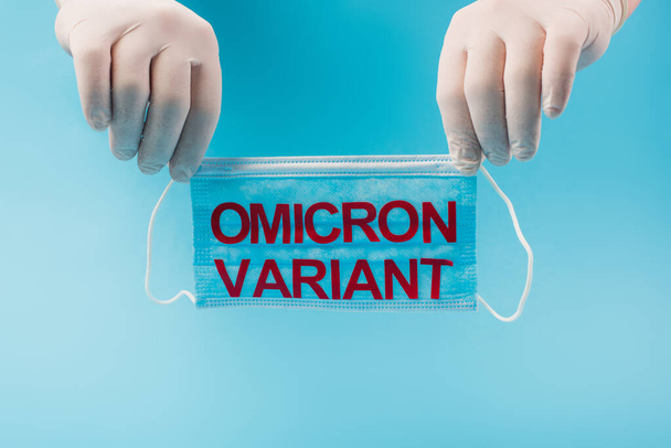 Cropped view of doctor in latex gloves holding medical mask with omicron variant lettering isolated on blue  - Foto, afbeelding