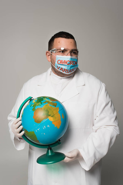 Scientist in medical mask with omicron variant lettering holding globe isolated on grey  - Fotó, kép