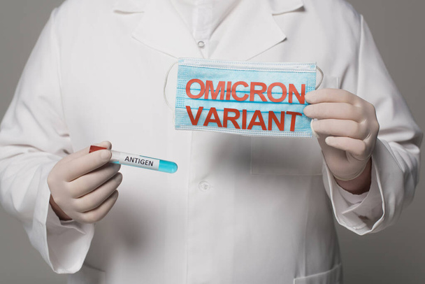 Cropped view of doctor holding medical mask with omicron variant and test tube with antigen lettering isolated on grey  - Fotó, kép