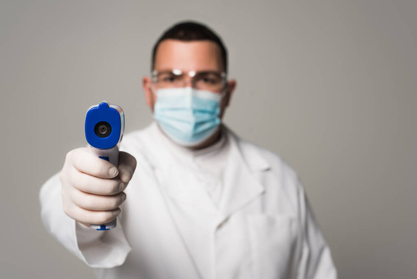 médico borroso en máscara médica sosteniendo pirómetro aislado en gris  - Foto, Imagen