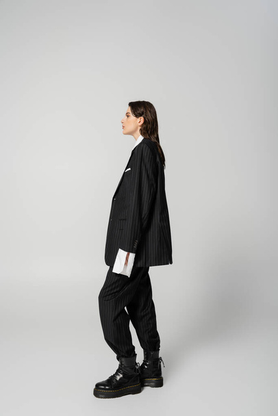 side view of trendy woman posing in black oversize pantsuit and rough boots on grey background - Foto, Bild