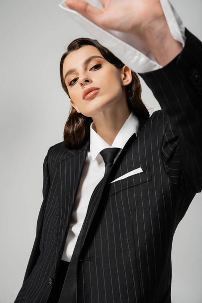 trendy woman in black striped blazer and white shirt with tie posing isolated on grey - Zdjęcie, obraz