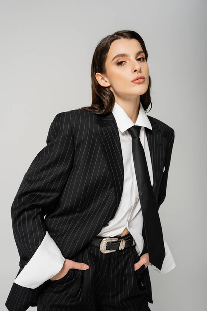 pretty young woman in black stylish suit and white shirt looking at camera while posing with hands in pockets isolated on grey - Foto, Imagem