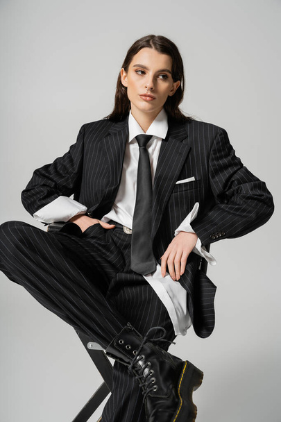 stylish brunette woman in elegant oversize suit sitting with hand in pocket while looking away isolated on grey - Foto, Bild