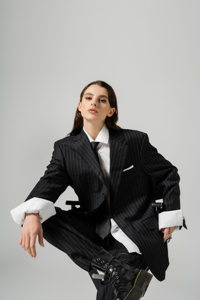 trendy woman in oversize formal wear sitting and looking at camera isolated on grey - Foto, afbeelding