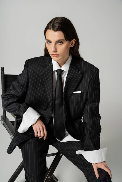 стильная брюнетка в черном полосатом костюме и галстуке позирует на стуле и смотрит на камеру, изолированную на сером - Фото, изображение