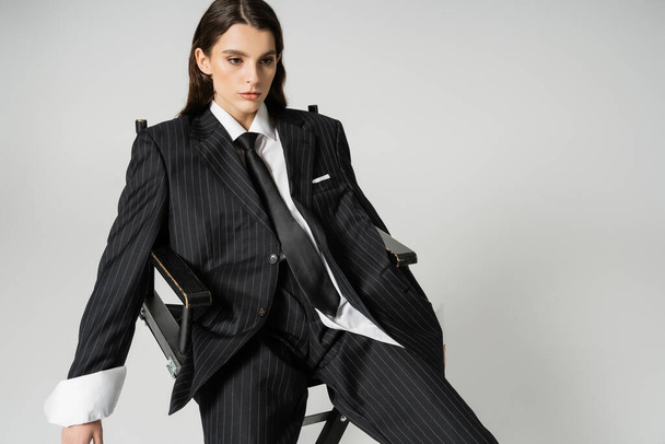 brunette woman in black striped pantsuit and tie sitting on chair isolated on grey - Zdjęcie, obraz