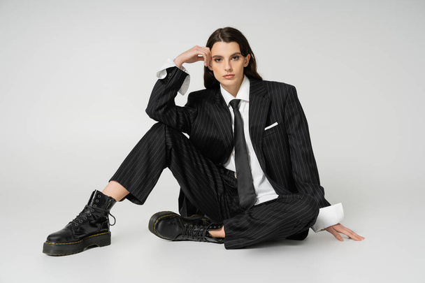 full length of young model in black elegant pantsuit and rough boots sitting with hand near head on grey background - Foto, Imagen