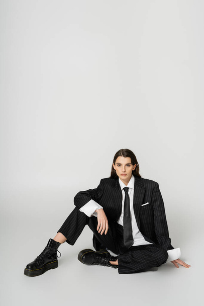 full length of fashionable woman in black elegant suit and rough boots sitting on grey background - Foto, afbeelding