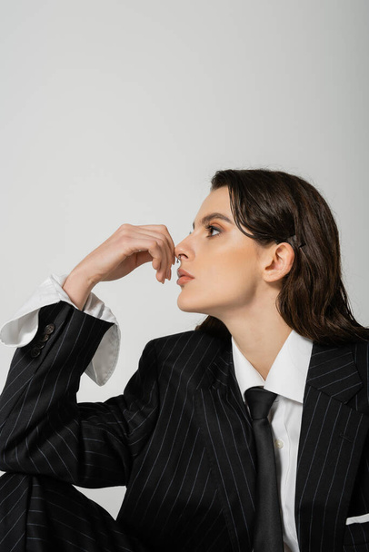 trendy woman in white shirt and black blazer with tie holding hand near face and looking away isolated on grey - Foto, Imagem