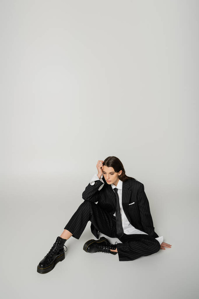 pretty woman in laced-up boots and oversize formal wear sitting with hand near head on grey background - Foto, afbeelding