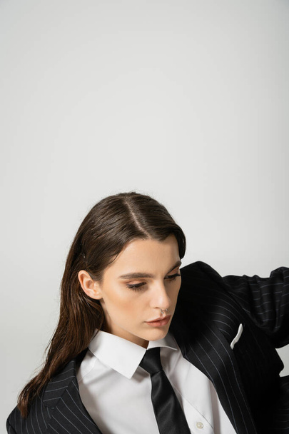brunette woman with makeup and piercing posing in black striped suit and tie isolated on grey - Foto, afbeelding