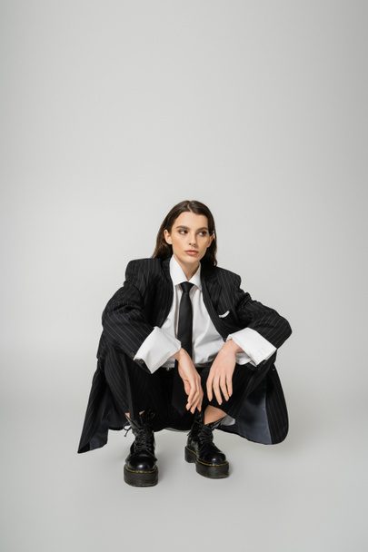 young model in black oversize suit and rough boots sitting on haunches and looking away on grey background - Foto, Imagen