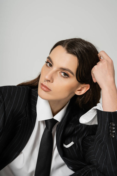 young woman with makeup and piercing wearing stylish formal wear and looking at camera isolated on grey - Foto, afbeelding
