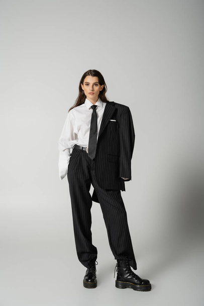 fashionable woman in oversize formal wear and laced-up boots standing with hand in pocket on grey background - Foto, afbeelding