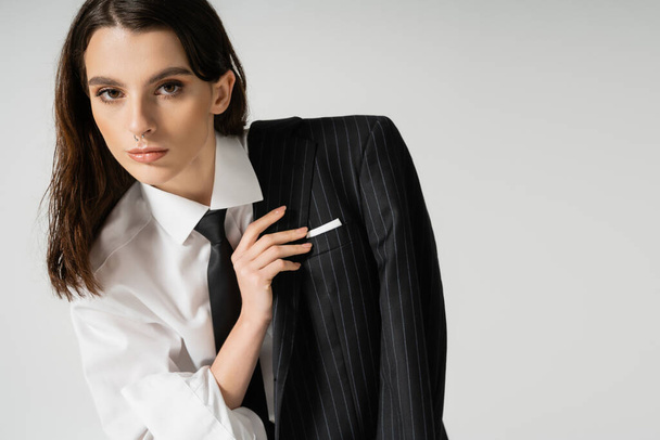 stylish woman with makeup and piercing wearing white shirt and holding black blazer isolated on grey - Foto, immagini
