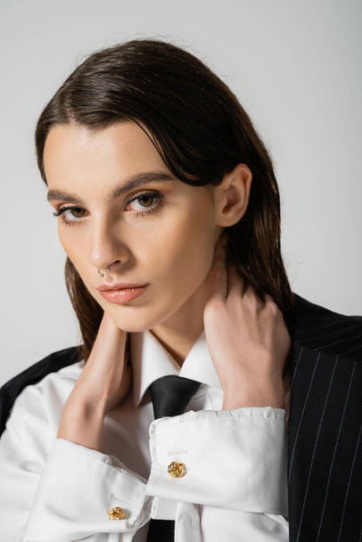 portrait of stylish woman in white shirt and black tie touching neck and looking at camera isolated on grey - Foto, Bild