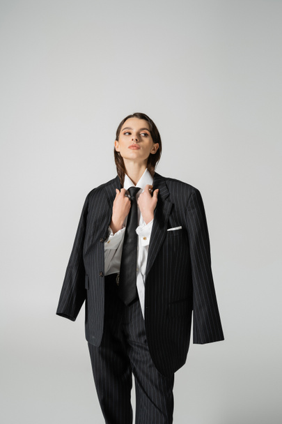 brunette woman in formal wear holding black oversize blazer and looking away isolated on grey - Zdjęcie, obraz