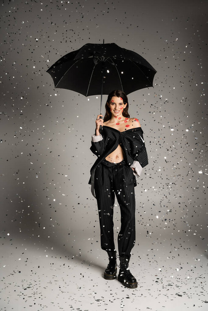 full length of smiling and stylish woman with red kisses on body standing with black umbrella under falling confetti on grey background - Photo, image